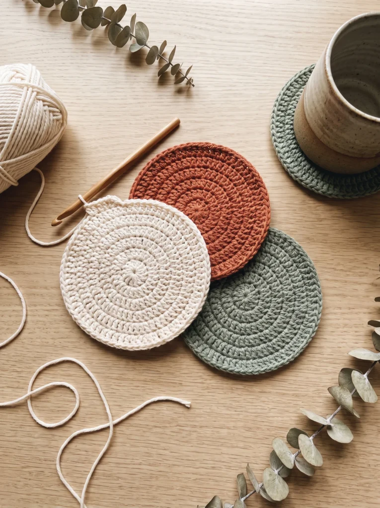 three round crochet coasters in cream terracotta and sage stacked near a stoneware mug, with a hook and yarn