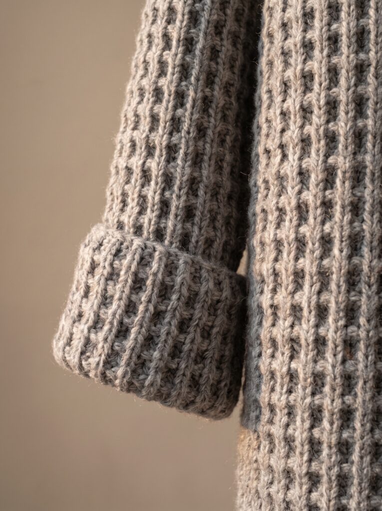 close-up of a gray crochet sweater sleeve showing a deep folded ribbed cuff against a taupe background