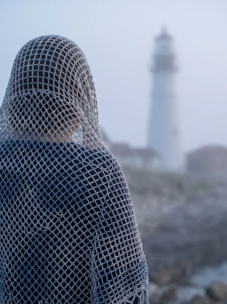 person wearing a grey blue open mesh crochet shawl over their head on a foggy coastline near a lighthouse