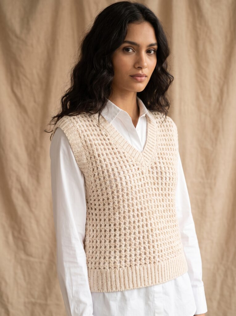 woman wearing a cream openwork crochet sweater vest layered over a white button down shirt against a linen backdrop