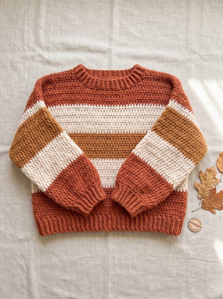 flat lay of a striped crochet sweater in terracotta mustard and cream folded on natural linen fabric with dried leaves