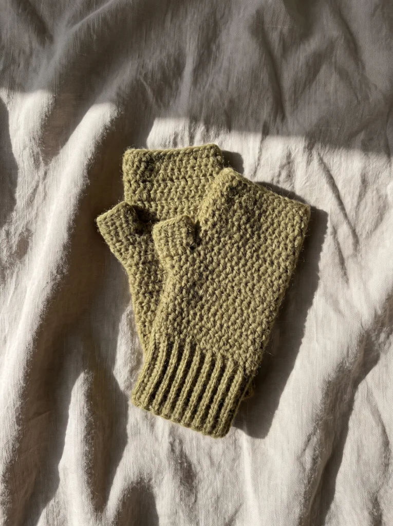 Pair of olive green fingerless crochet mitts folded on a wrinkled linen cloth