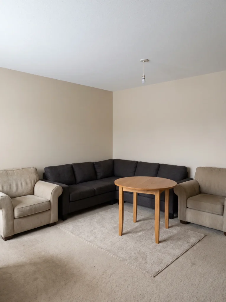 bare beige carpeted apartment living room with a dark sectional, two tan armchairs, a small round oak table, and a bare ceiling bulb